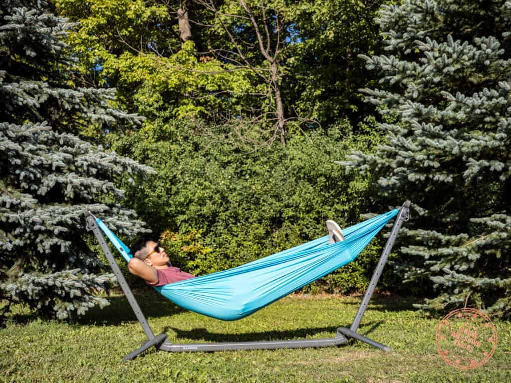 man sitting in a kammok roo single attached to a swiftlet portable hammock stand while in the park near trees